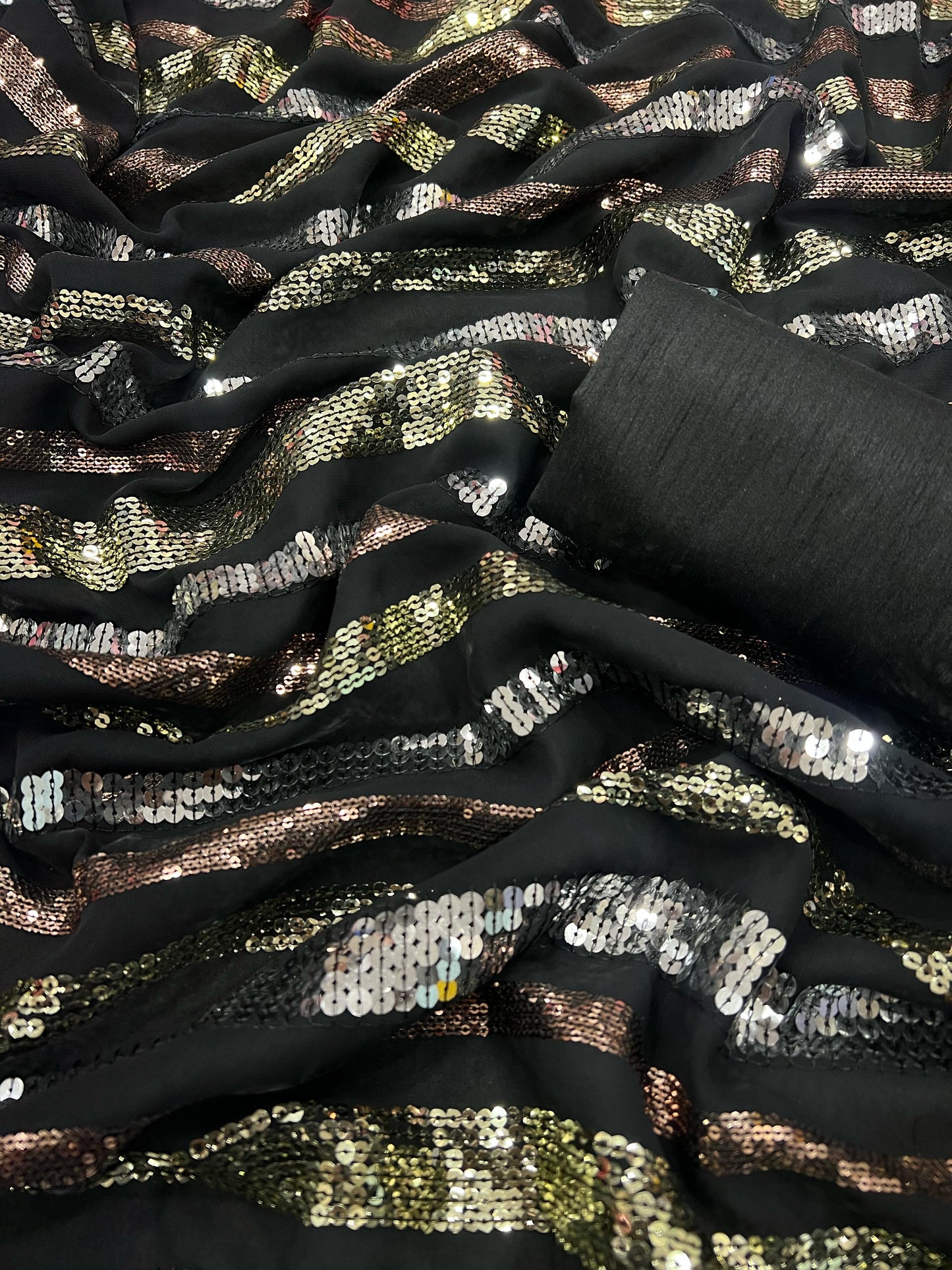 The Black Georgette Saree, perfect for crafting a dazzling lehenga, is adorned with sequins in silver, gold, and bronze tones. A roll of this shimmering fabric can be seen partially in the upper right corner.