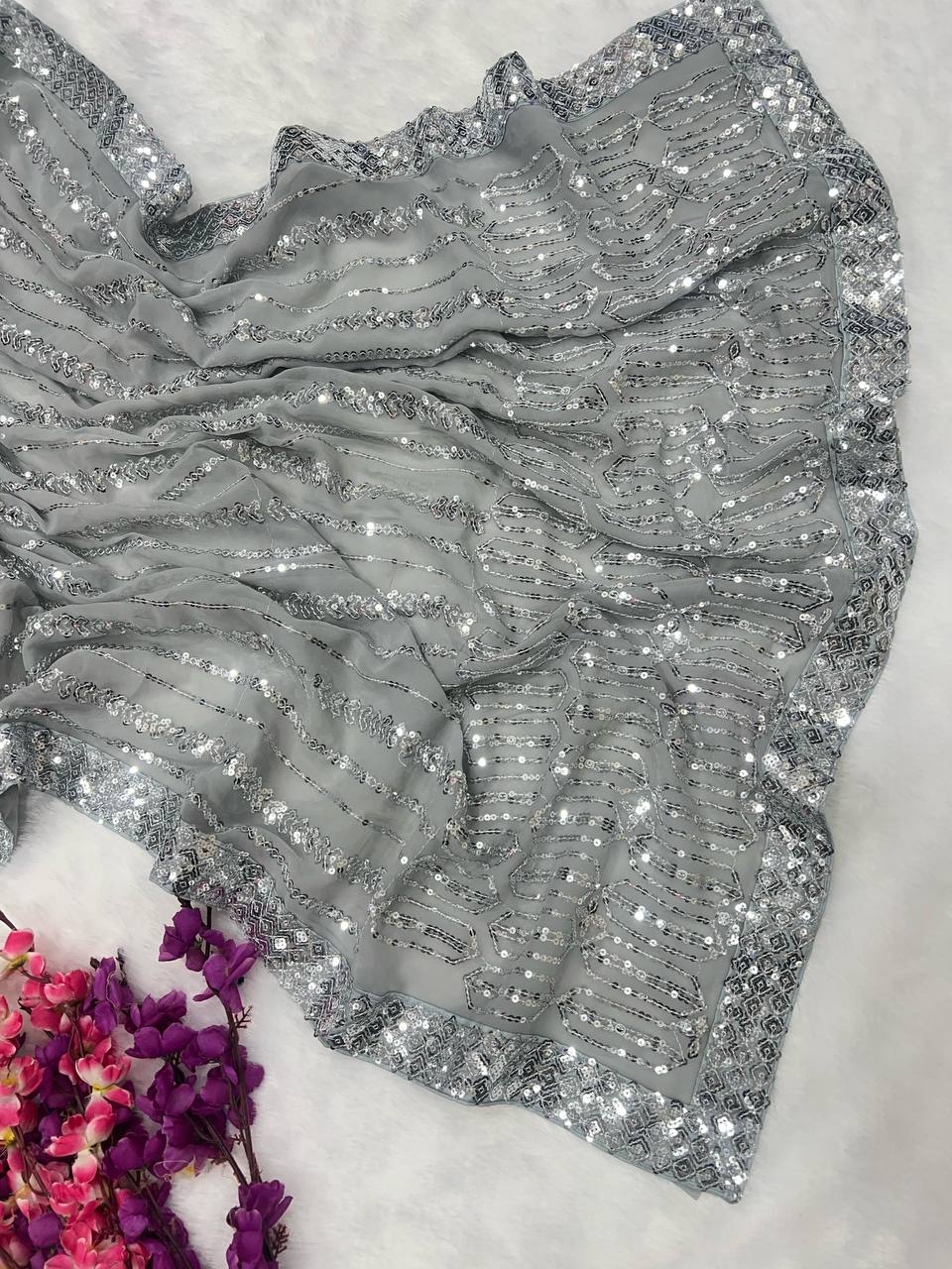 The Pastel Grey Bollywood Style Fully Sequins Saree with Stitched Blouse drapes elegantly, showcasing an ornate lehenga-like border and a cluster of vibrant purple and pink flowers on a white surface.