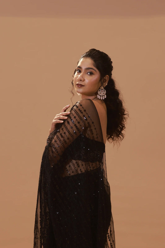 A woman with long, curly hair stands against a beige background in a Full Black Net Sequins Saree With Stitched Blouse and large ornate earrings, smiling slightly over her shoulder with elegance similar to an embroidered kurta, one hand gently touching her upper arm.