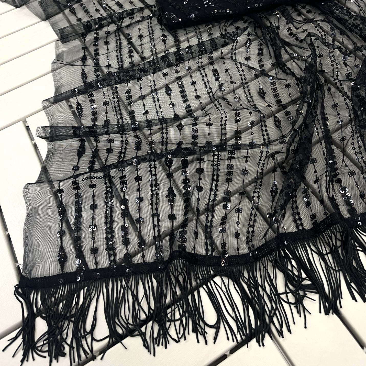 The Full Black Net Sequins Saree With Stitched Blouse, adorned with sequins in vertical and geometric patterns, rests elegantly on a white wooden surface, accompanied by a sheer black scarf with fringe trim.
