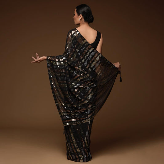 A woman poses against a brown backdrop in a Black Georgette Saree with Multi Colour Sequins work and Stitched Blouse. Facing away, she showcases the sarees intricate patterns reminiscent of a traditional sharara, her hair styled in a neat bun.