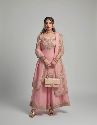Woman in a pink traditional outfit standing on a checkered floor with plants and vehicles in the background.