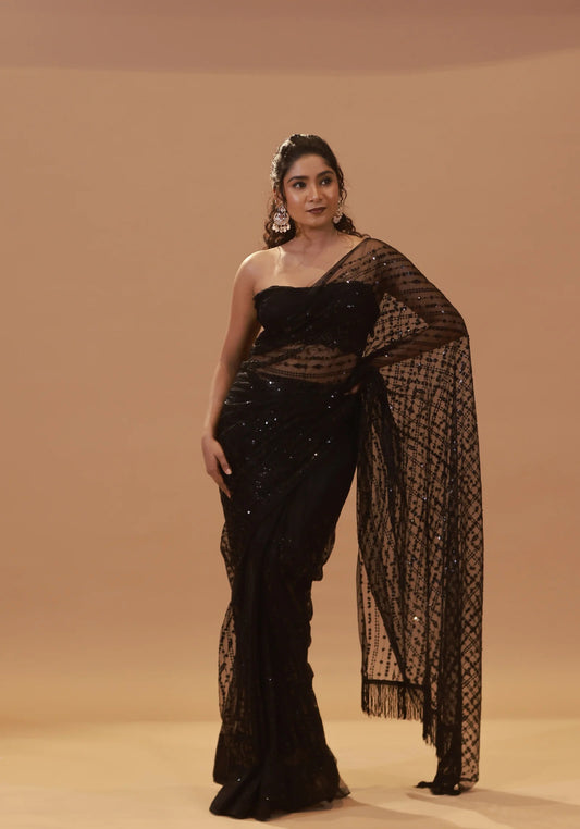 A woman models a Full Black Net Sequins Saree With Stitched Blouse in front of a plain backdrop. She stands sideways, hand on hip, with the saree draped over her arm. Her updo is accentuated by hoop earrings that shimmer like embellishments on a choli.
