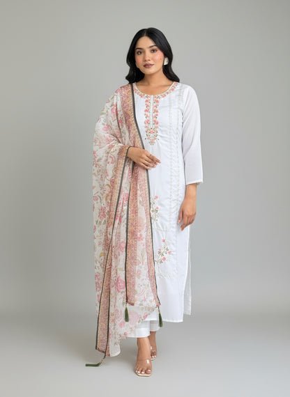 Woman in a white and pink embroidered dress standing in a room with a mirror and decorative rug.