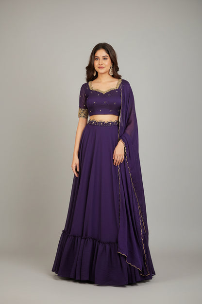 Woman in a purple traditional outfit with gold details standing in a decorated room.