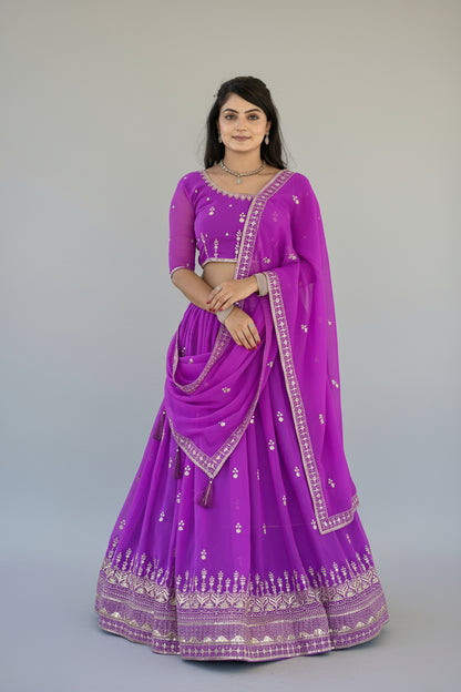 Woman wearing a vibrant purple traditional outfit with intricate designs, standing against a decorative wall.