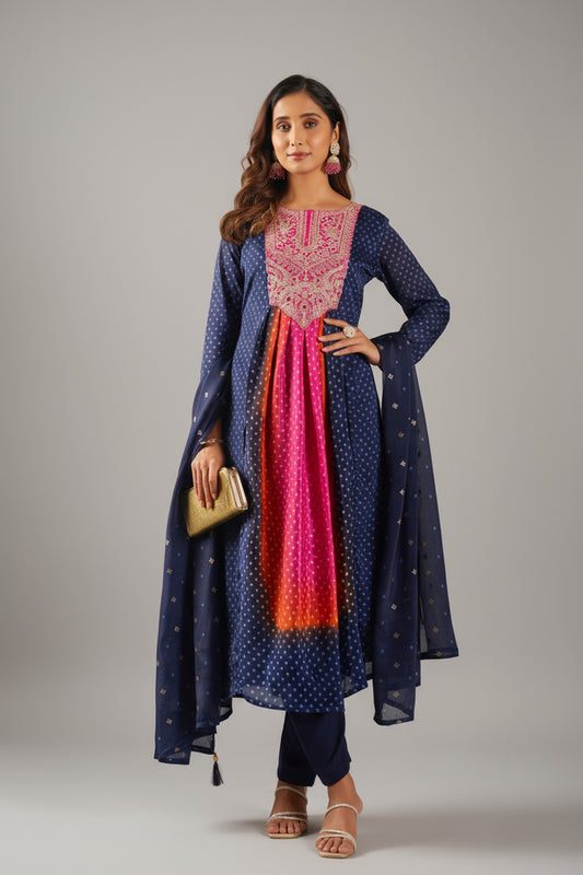 Woman in a traditional outfit with a blue and pink dress and orange dupatta standing indoors.