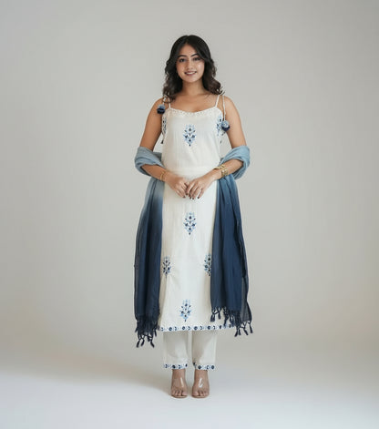 Woman in a white outfit with blue accents standing indoors next to a vase of flowers.